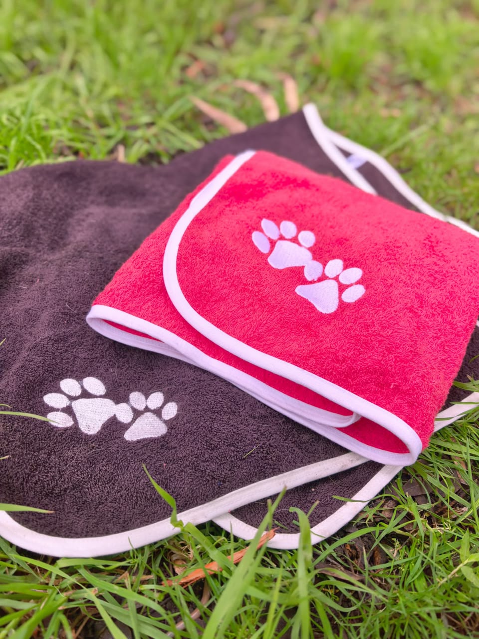 Toallas Pet Towels café y roja sobre pasto, con bordados de huellas y ribetes claros